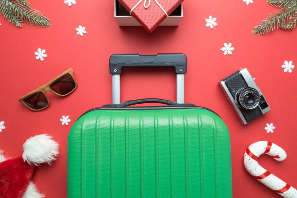 Best holiday gifts for travelers: Creative composition made of suitcase, gift box, sunglasses, santa hat, candy cane, retro photo camera, fir branches and snowflakes on red background.