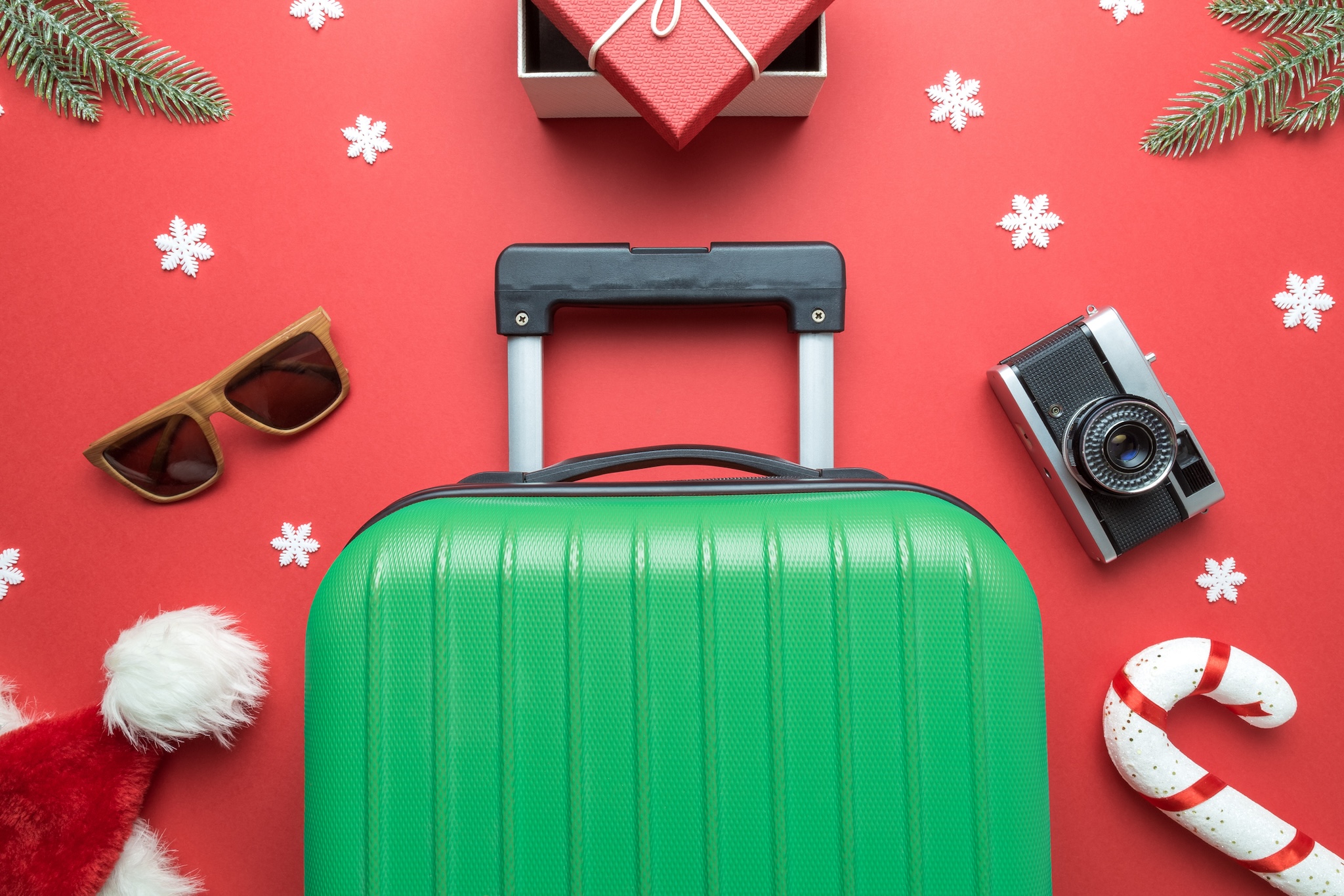 Best holiday gifts for travelers: Creative composition made of suitcase, gift box, sunglasses, santa hat, candy cane, retro photo camera, fir branches and snowflakes on red background.